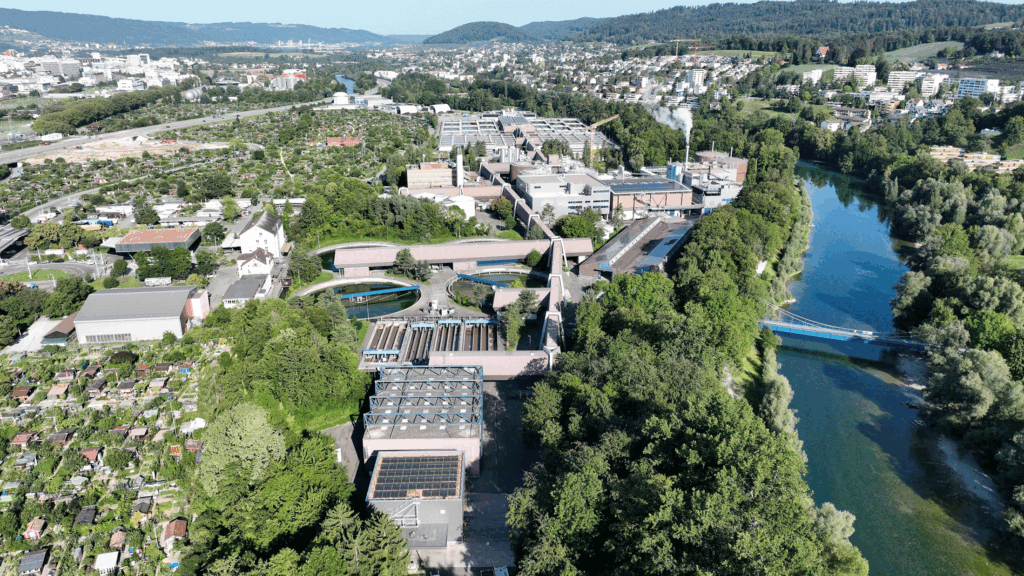 Luftaufnahme eines Industriekomplexes neben einem Fluss mit umliegenden Wohngebieten und Grünflächen.
