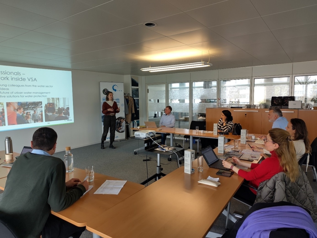 Eine Gruppe von Menschen h&ouml;rt sich in einem Konferenzraum eine Pr&auml;sentation zum Thema st&auml;dtisches Wassermanagement an. Ein Vortragender steht neben einem Bildschirm mit Bildern. Die Tische sind U-f&ouml;rmig angeordnet.