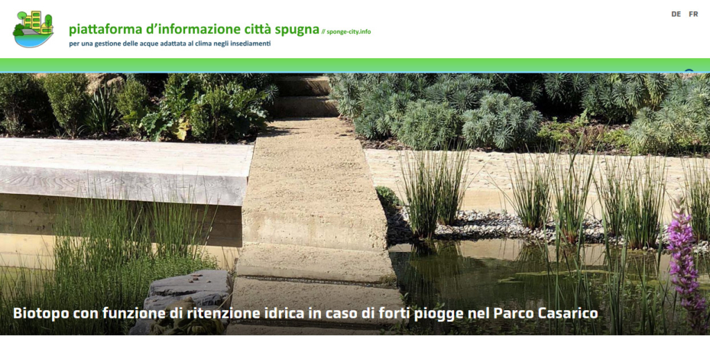 Ein Steinweg über einem Rückhaltebecken, umgeben von viel Grün im Parco Casarico. Der Text lautet: „Biotopo con funzione di ritenzione idrica in caso di forti piogge nel Parco Casarico.“
