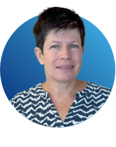 Eine Frau mit kurzen dunklen Haaren und einer gemusterten Bluse steht vor einem kreisförmigen Hintergrund mit blauem Farbverlauf, schaut in die Kamera und lächelt leicht.