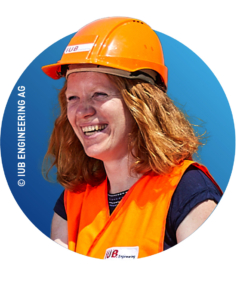 Eine Person mit roten Haaren lächelt, während sie einen orangefarbenen Schutzhelm und eine Warnweste vor einem blauen Hintergrund trägt.