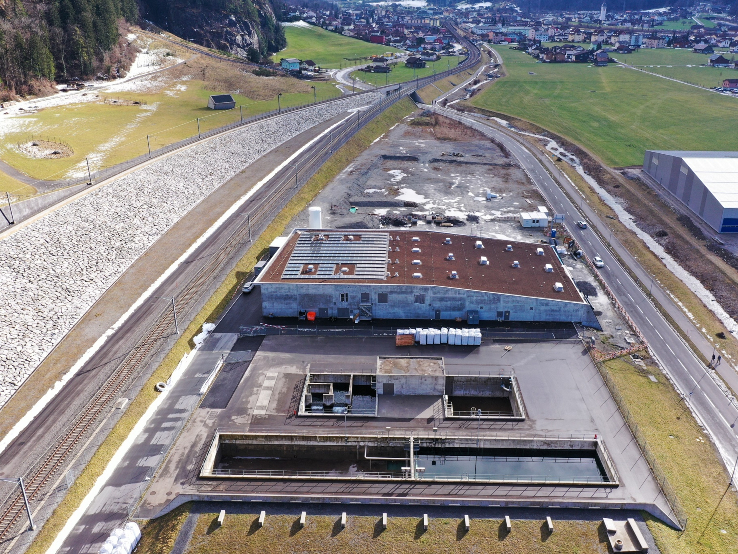 Luftaufnahme einer modernen Wasseraufbereitungsanlage mit einem rechteckigen Hauptgeb&auml;ude, offenen Wassertanks und umliegenden Stra&szlig;en und Bahngleisen in einem l&auml;ndlichen Gebiet.
