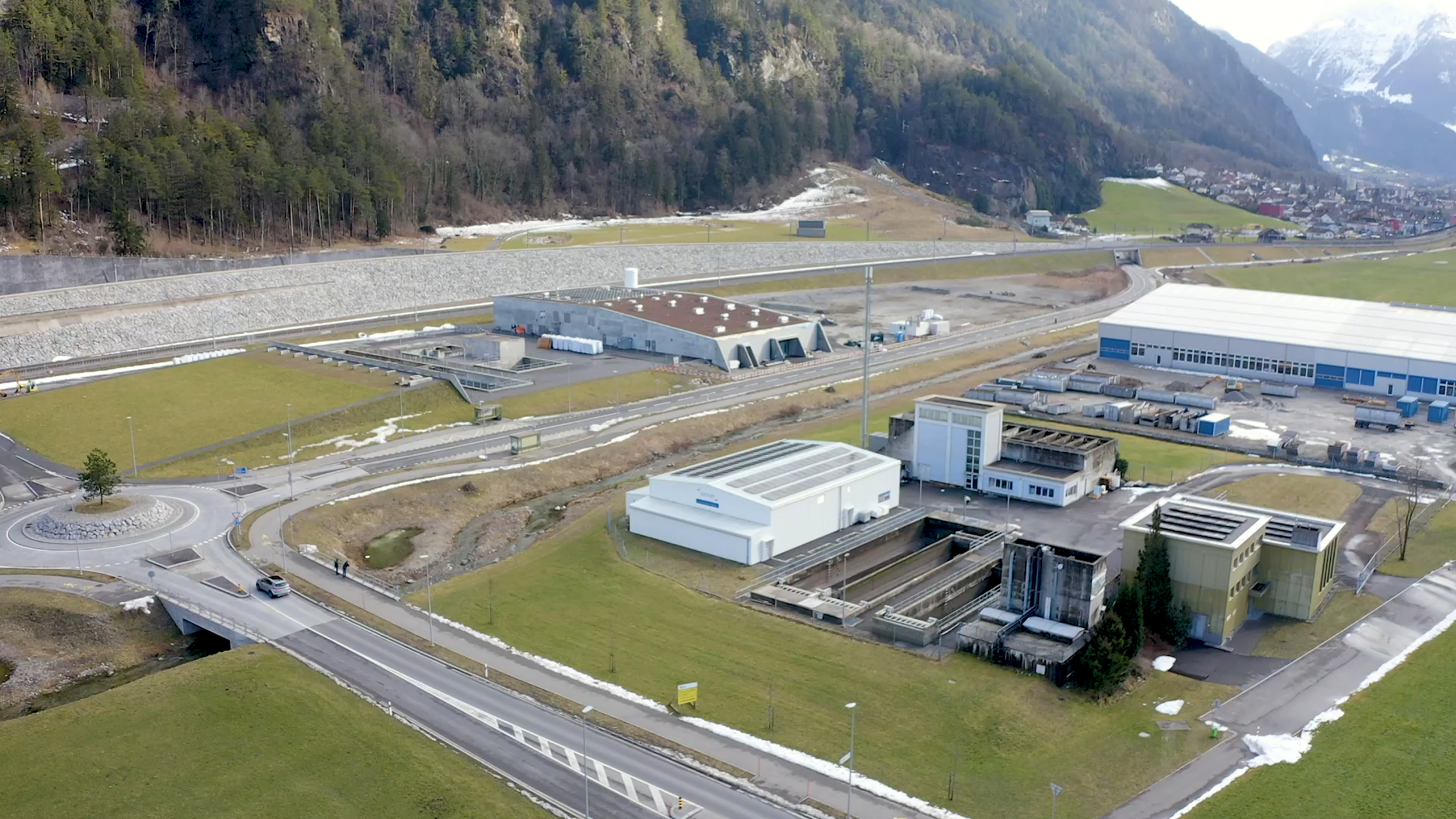 Luftaufnahme eines Industriekomplexes mit mehreren Geb&auml;uden, Stra&szlig;en und Gr&uuml;nfl&auml;chen in der N&auml;he von bewaldeten H&uuml;geln und einer absch&uuml;ssigen B&ouml;schung.