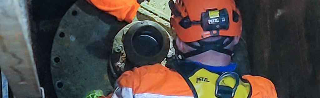 Ein Arbeiter mit orangefarbener Schutzkleidung und Helm inspiziert oder wartet Industriemaschinen in einem geschlossenen Raum.