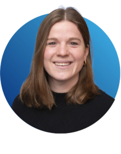 Eine Frau mit glattem braunem Haar, die ein schwarzes Oberteil und goldene Ohrringe tr&auml;gt, l&auml;chelt vor einem blauen runden Hintergrund.