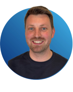 Ein Mann mit kurzen braunen Haaren und einem Bart, der ein dunkles Hemd tr&auml;gt, l&auml;chelt in die Kamera vor einem blauen, runden Hintergrund.