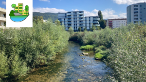 Ein klarer Bach flie&szlig;t durch dichtes gr&uuml;nes Geb&uuml;sch mit modernen Wohngeb&auml;uden im Hintergrund unter einem teilweise bew&ouml;lkten Himmel; ein Stadtbildsymbol erscheint in der oberen linken Ecke.