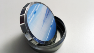 Ein runder Metallbeh&auml;lter mit einem blauen Etikett auf der Innenseite des Deckels, auf dem ein deutscher Text und ein Bild von Wasser abgebildet sind, steht ge&ouml;ffnet auf einer wei&szlig;en Fl&auml;che.