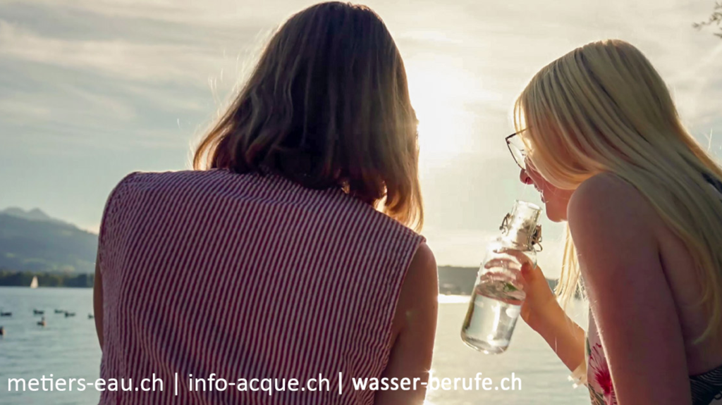 Zwei Frauen sitzen an einem See, eine trinkt Wasser aus einer Flasche, im Hintergrund schwimmen Enten und Berge. Das Sonnenlicht ist sichtbar. Links zur Website werden am unteren Rand des Bildes angezeigt.