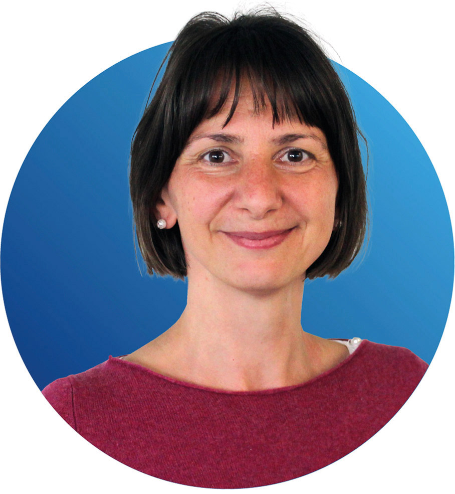 Eine Frau mit kurzen dunklen Haaren, die ein weinrotes Oberteil und Ohrstecker trägt und vor einem blauen, runden Hintergrund lächelt.