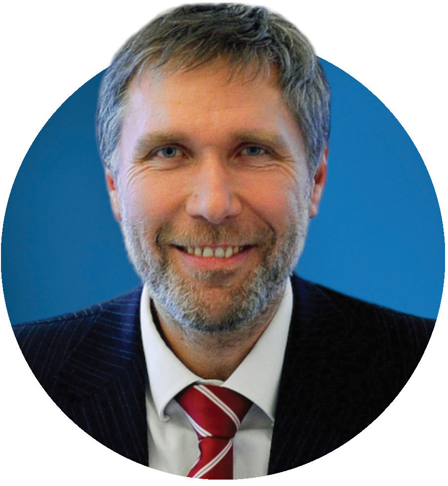 Ein Mann mit kurzen grauen Haaren und Bart, der einen dunklen Nadelstreifenanzug, ein wei&szlig;es Hemd und eine rot gestreifte Krawatte tr&auml;gt, l&auml;chelt vor einem blauen Hintergrund.