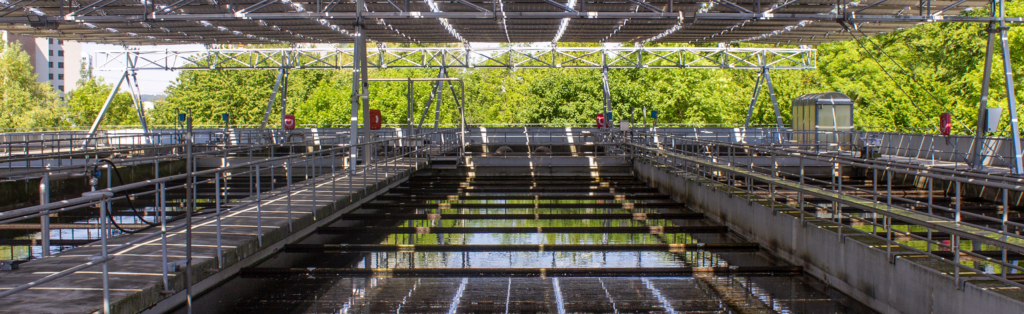 Reihen von Betonbecken mit Metallgeländern und Überbauungen in einer Wasseraufbereitungsanlage, umgeben von Bäumen.
