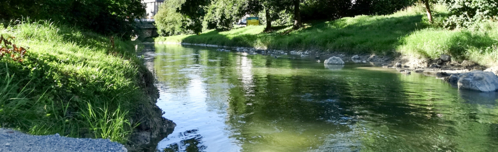 Ein ruhiger Fluss fließt zwischen grasbewachsenen Ufern, die auf beiden Seiten von Bäumen gesäumt sind, und im Hintergrund ist bei strahlendem Tageslicht eine Steinbrücke zu sehen.